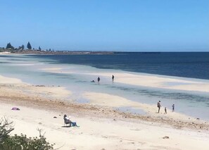 A beach with clear water, people walking, and a person sitting on a chair.