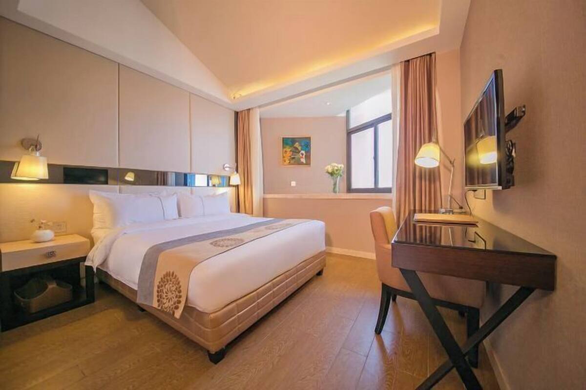 A hotel room with a bed, bedside table, desk, chair, TV, and a painting on the wall.