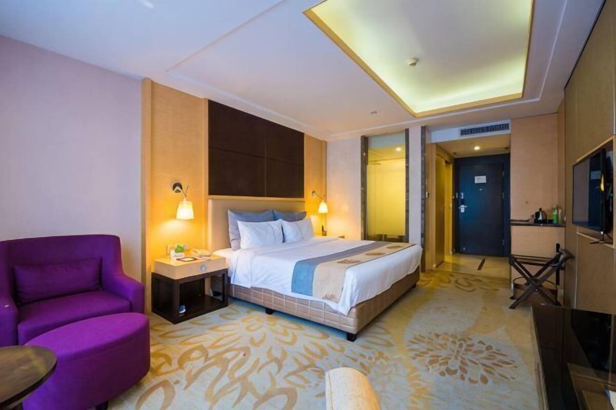 A hotel room with a bed, a purple armchair, a TV, and a desk with a lamp.