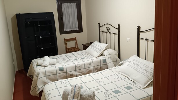 A bedroom with two beds, a wardrobe, a chair, and a window with a curtain. - Casa Rural Abuela Maria (Pasarón de la Vera)