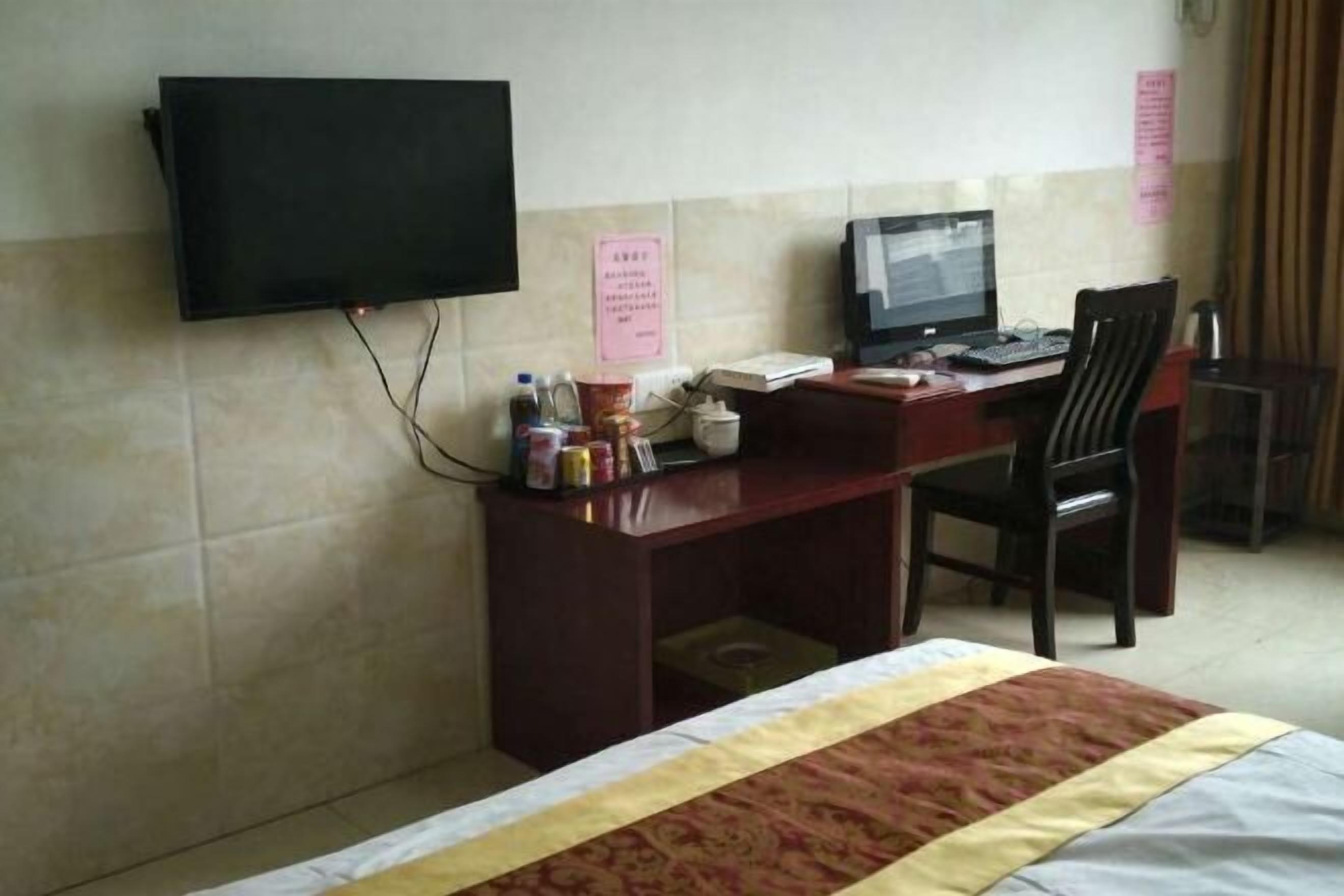 A hotel room with a bed, a desk with a computer, a chair, a television mounted on the wall, and a small table with various items.