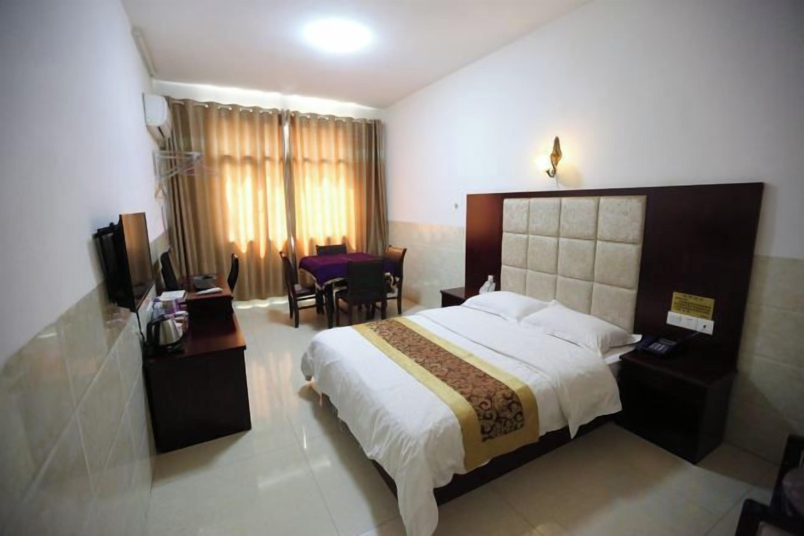 A hotel room with a bed, bedside table, desk, chair, and a view of the outdoors through curtains.