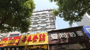 Vista de la calle con varios comercios, incluyendo un letrero amarillo con texto rojo, un escaparate negro con texto blanco y un edificio con múltiples ventanas al fondo.
