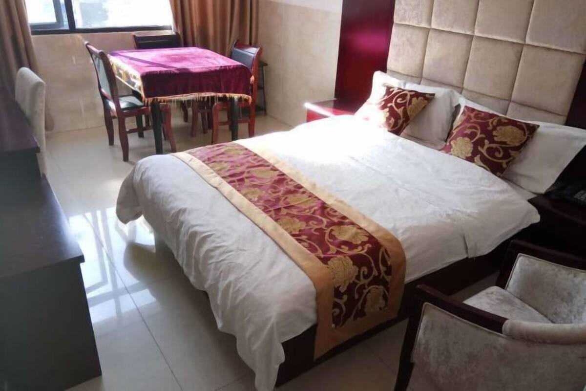 A hotel room with a large bed, a dining table with chairs, and a view of the outdoors through a window.
