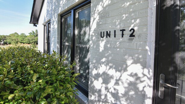 Un bâtiment à l’extérieur en briques blanches, avec une porte portant l’inscription « UNITÉ 2 » et une fenêtre.