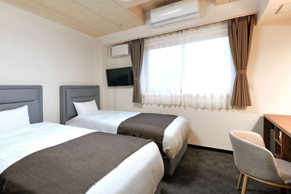Habitación de hotel con dos camas, un escritorio, una silla, un equipo de aire acondicionado y un televisor.