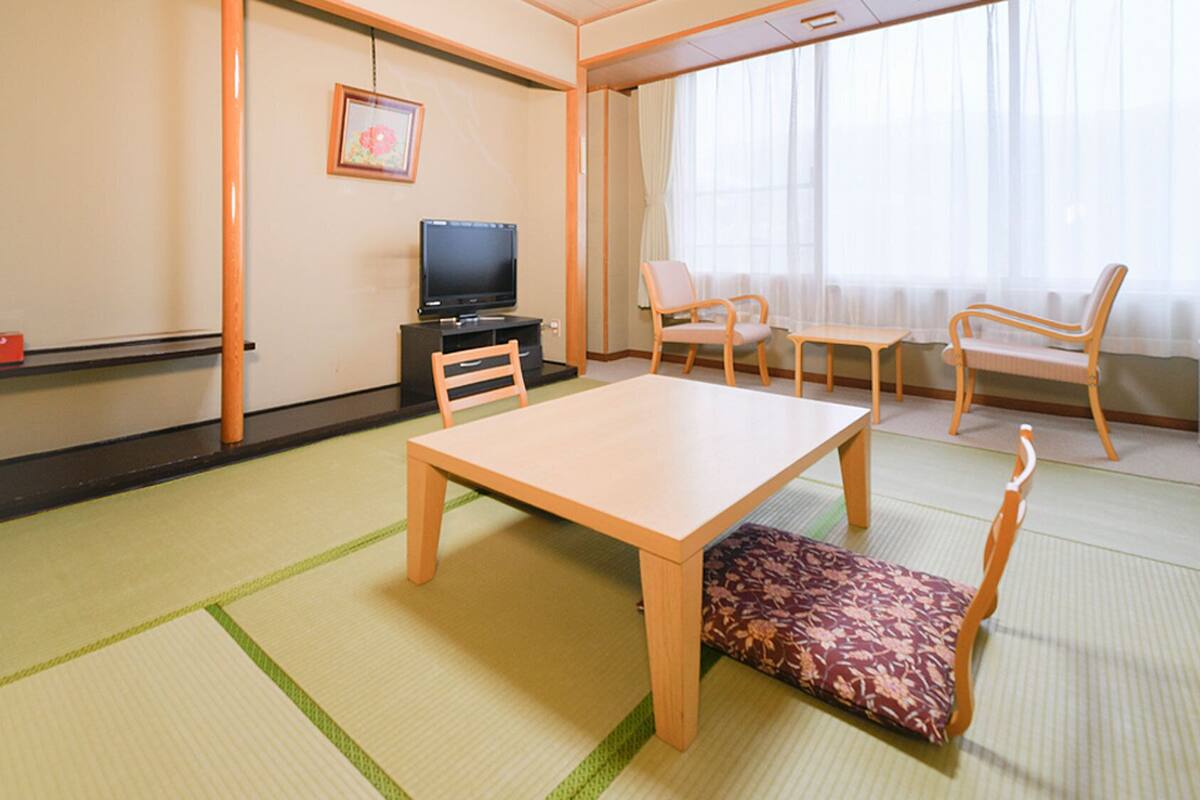 Una habitación tradicional japonesa con suelo de tatami, una mesa baja, sillas, un televisor y un cuadro en la pared.
