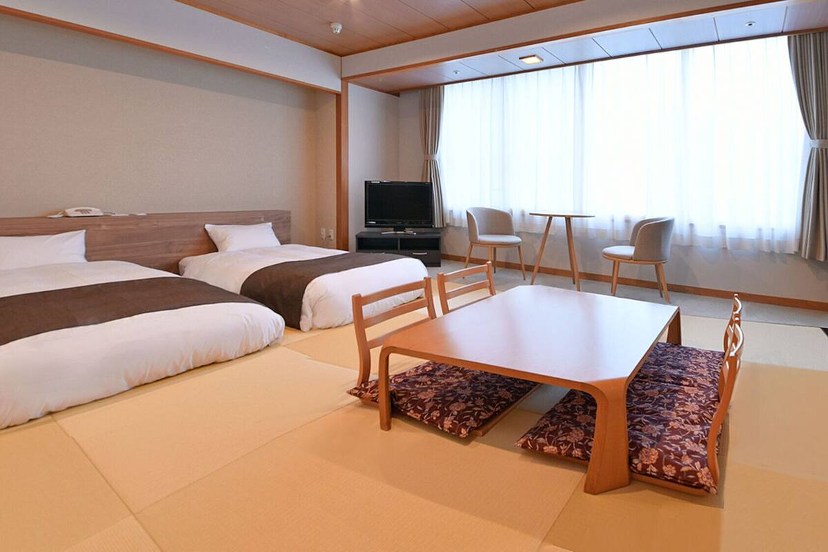 Habitación de hotel con dos camas, televisión, mesa de comedor y vistas al exterior.
