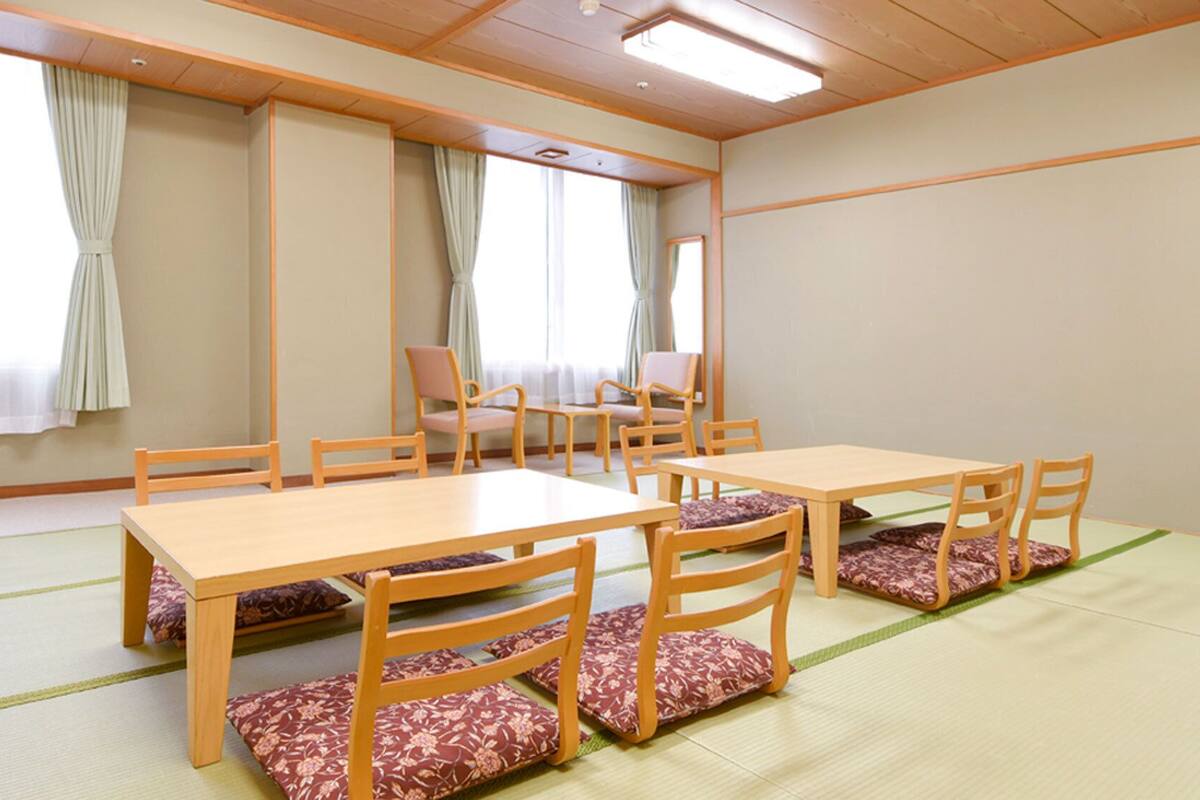 Una habitación con tatamis, mesas y sillas de madera, una ventana grande y cortinas.