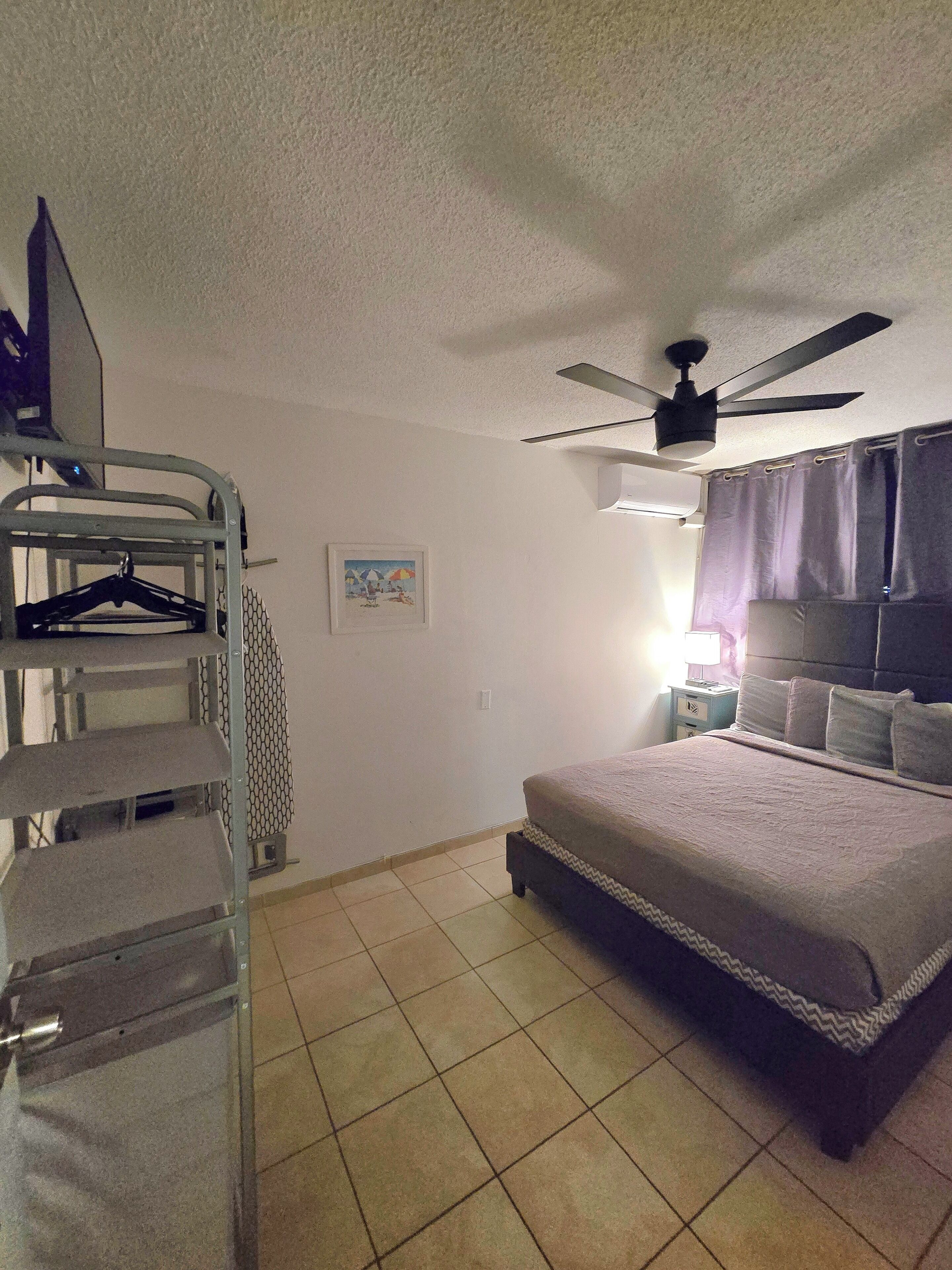 A bedroom with a bed, a ceiling fan, a wall-mounted heater, and a metal shelf unit.