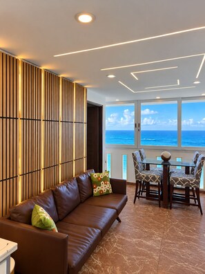 A modern living room with a leather sofa, patterned cushions, a dining table with chairs, and a view of the ocean through large windows.
