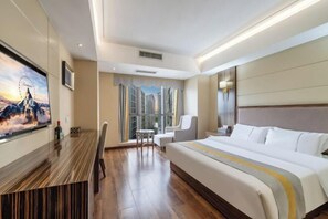 Quarto de hotel com cama grande, escrivaninha com televisão, área de refeições e vista para a cidade através de amplas janelas.