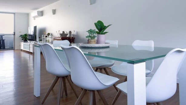 A modern dining area with a glass table and white chairs, a potted plant, and a kitchen in the background.