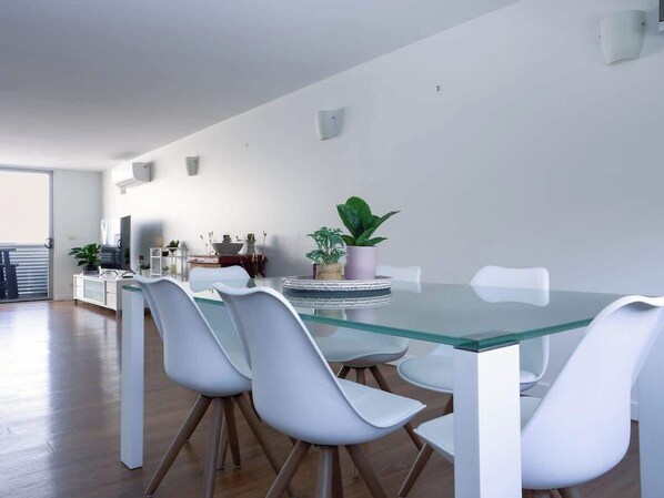 A modern dining area with a glass table and white chairs, a potted plant, and a kitchen in the background.