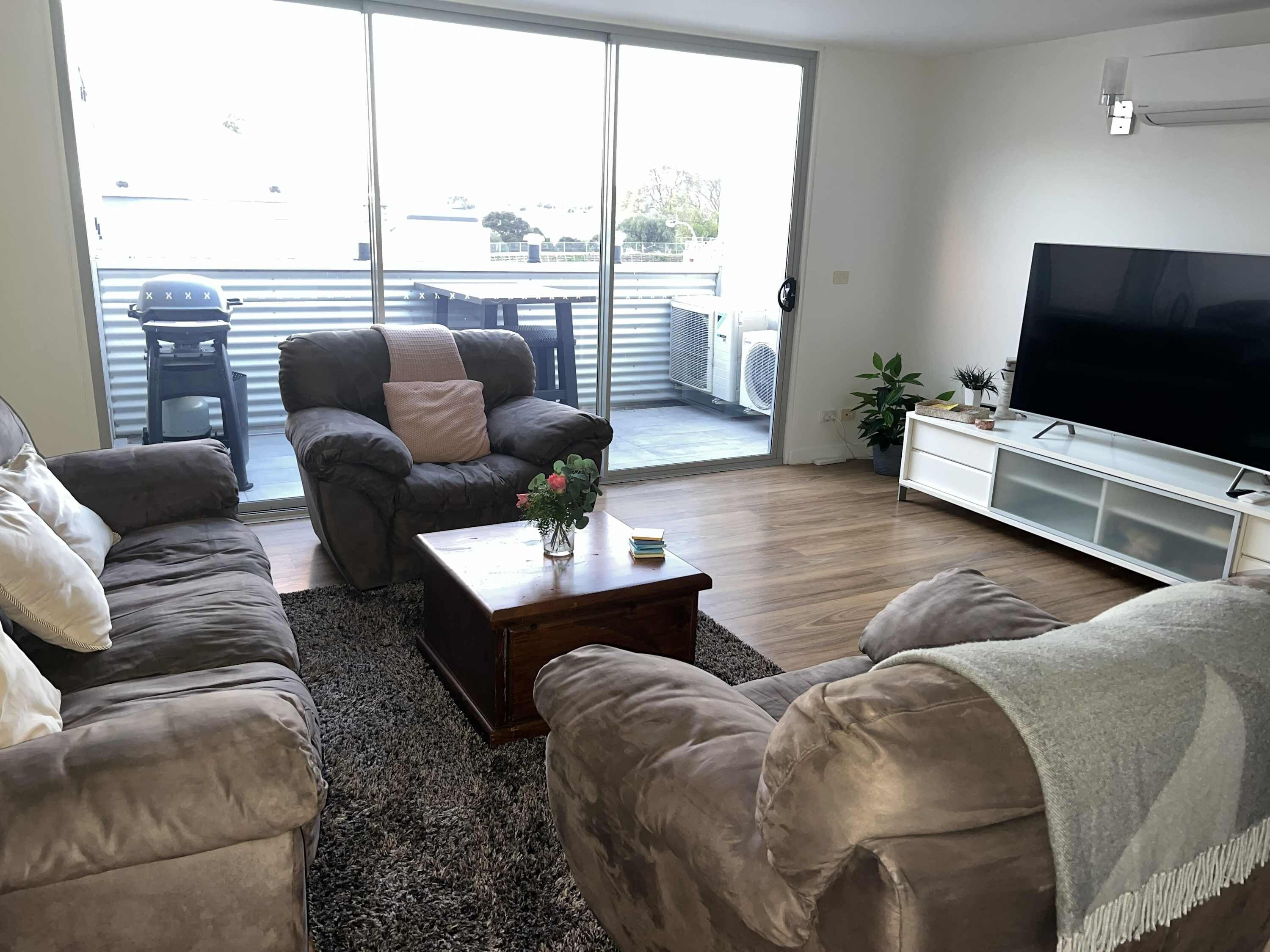 A living room with a sofa set, a coffee table with a vase, a flat-screen TV, and a sliding glass door leading to an outdoor area with a patio table and chairs.