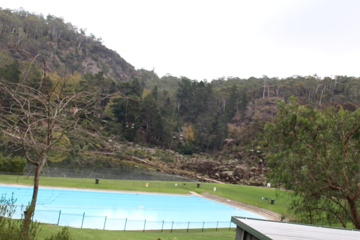 A swimming pool with a surrounding grassy area, a building with a sloped roof, and a forested hillside in the background.