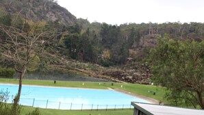 A swimming pool with a surrounding grassy area, a building with a sloped roof, and a forested hillside in the background.