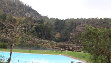 A swimming pool with a surrounding grassy area, a building with a sloped roof, and a forested hillside in the background.