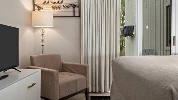 Quarto de hotel com cama, poltrona, televisão sobre uma cômoda, luminária e janela com cortinas.
