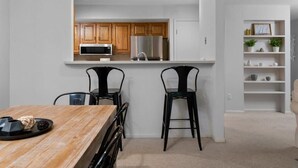 A modern kitchen with wooden cabinets, a stainless steel refrigerator, and a microwave. There is a dining area with a wooden table and two black chairs. - GT Resort Area I Fireplace + Golf Views (Williamsburg)