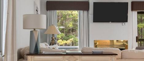 A hotel room with a bed, a desk with a lamp, a TV, and a view of greenery outside.