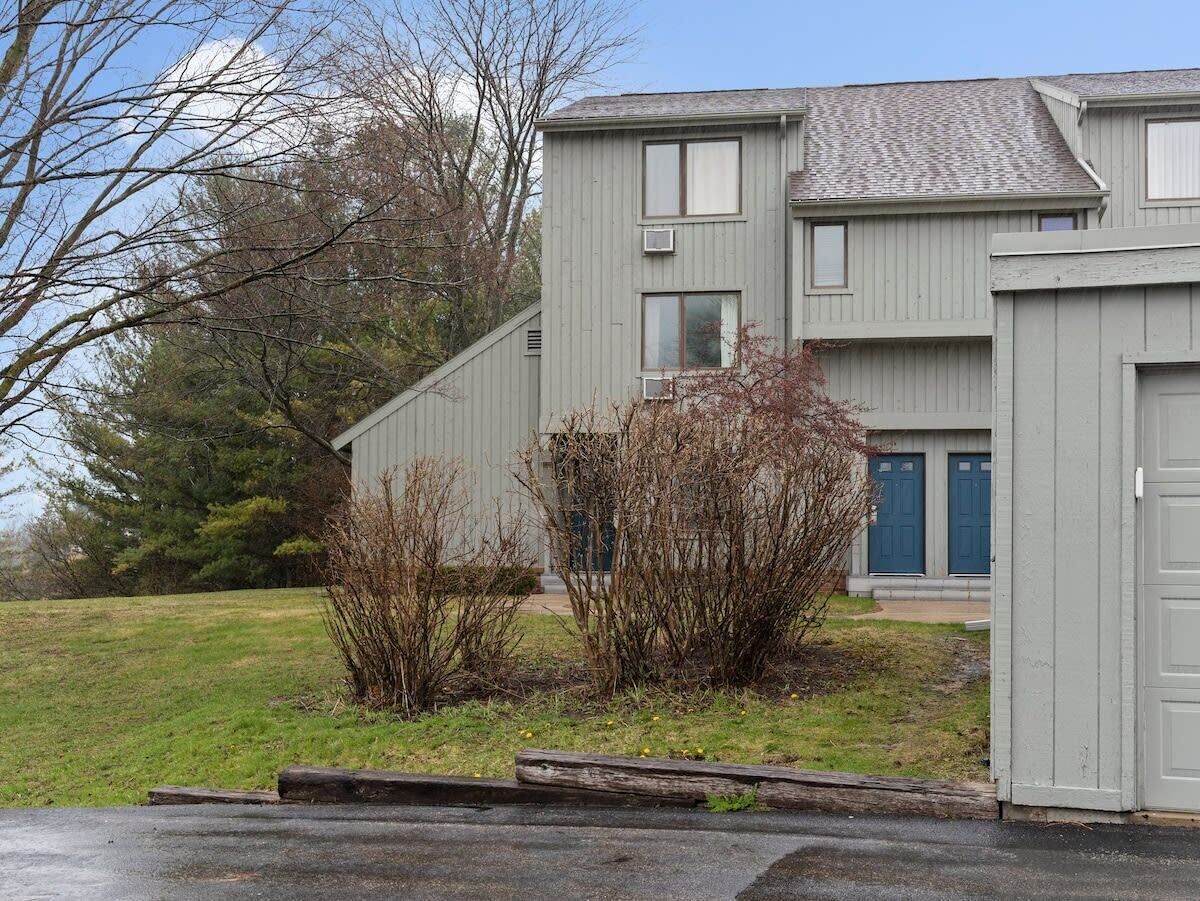 A two-story building with a flat roof, a garage on the right, and a small front yard with shrubs and a tree.