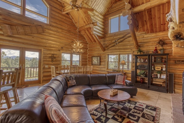A log cabin interior with a large leather sectional sofa, a round coffee table, and a wooden bookshelf.