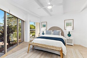 Ein geräumiges Schlafzimmer mit einem großen Bett, einem Deckenventilator und Blick auf ein Gewässer durch große Fenster.