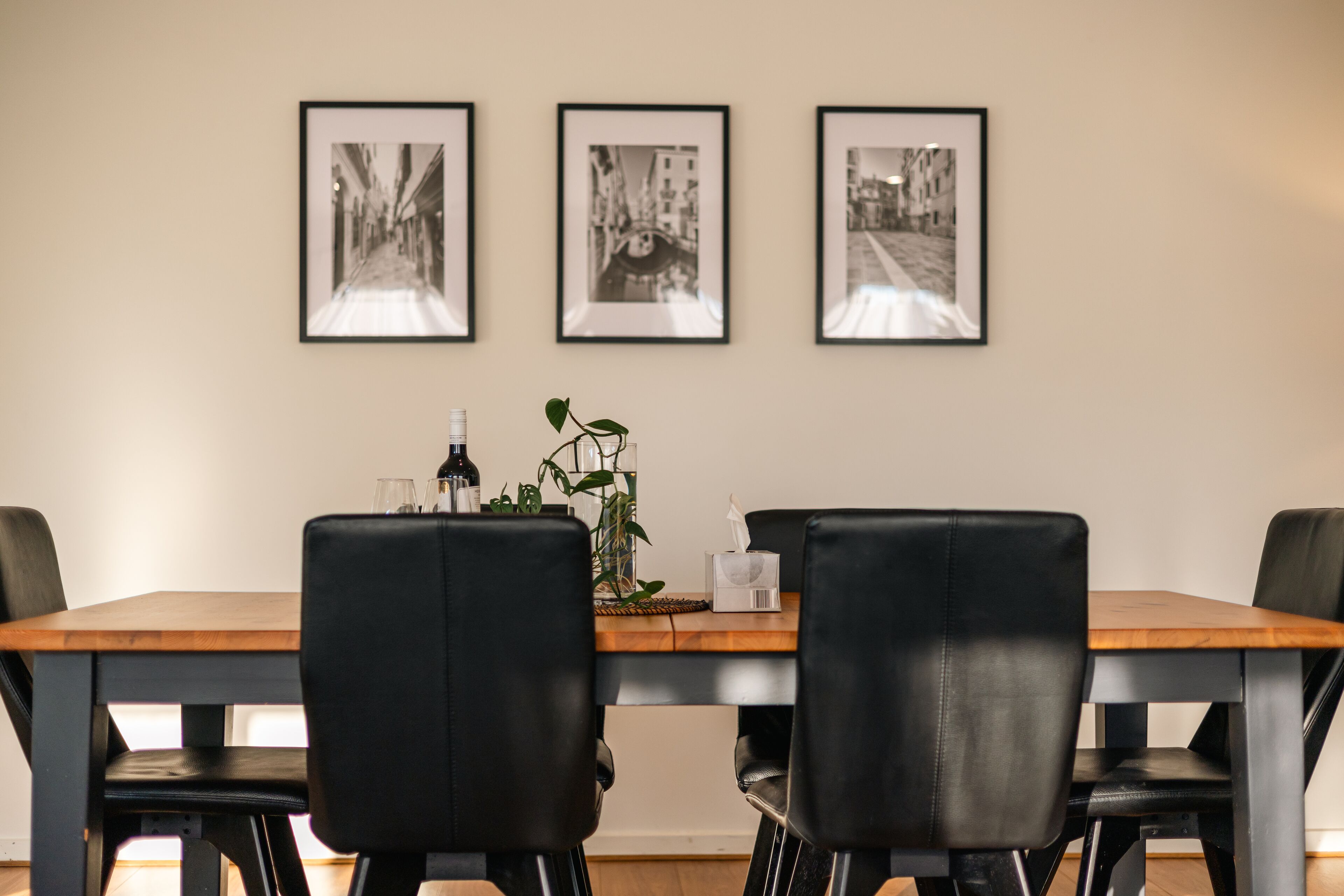 A dining area with a wooden table, black chairs, and three framed black and white photographs on the wall.