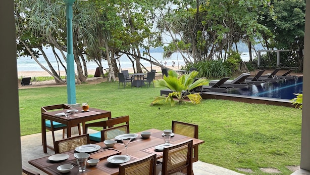 A covered outdoor dining area with tables and chairs, a pool, and a view of the beach and ocean. - Villa Talay (Talalla)