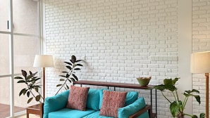 A room with a white brick wall, a wooden sofa with teal cushions, a wooden coffee table, a ceiling fan, and a large window. - Villa Talay (Talalla)