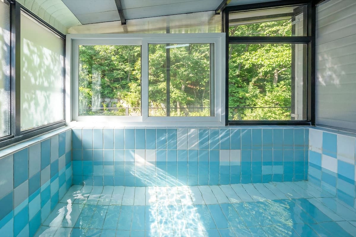 A swimming pool with blue tiles and large windows overlooking a green landscape.