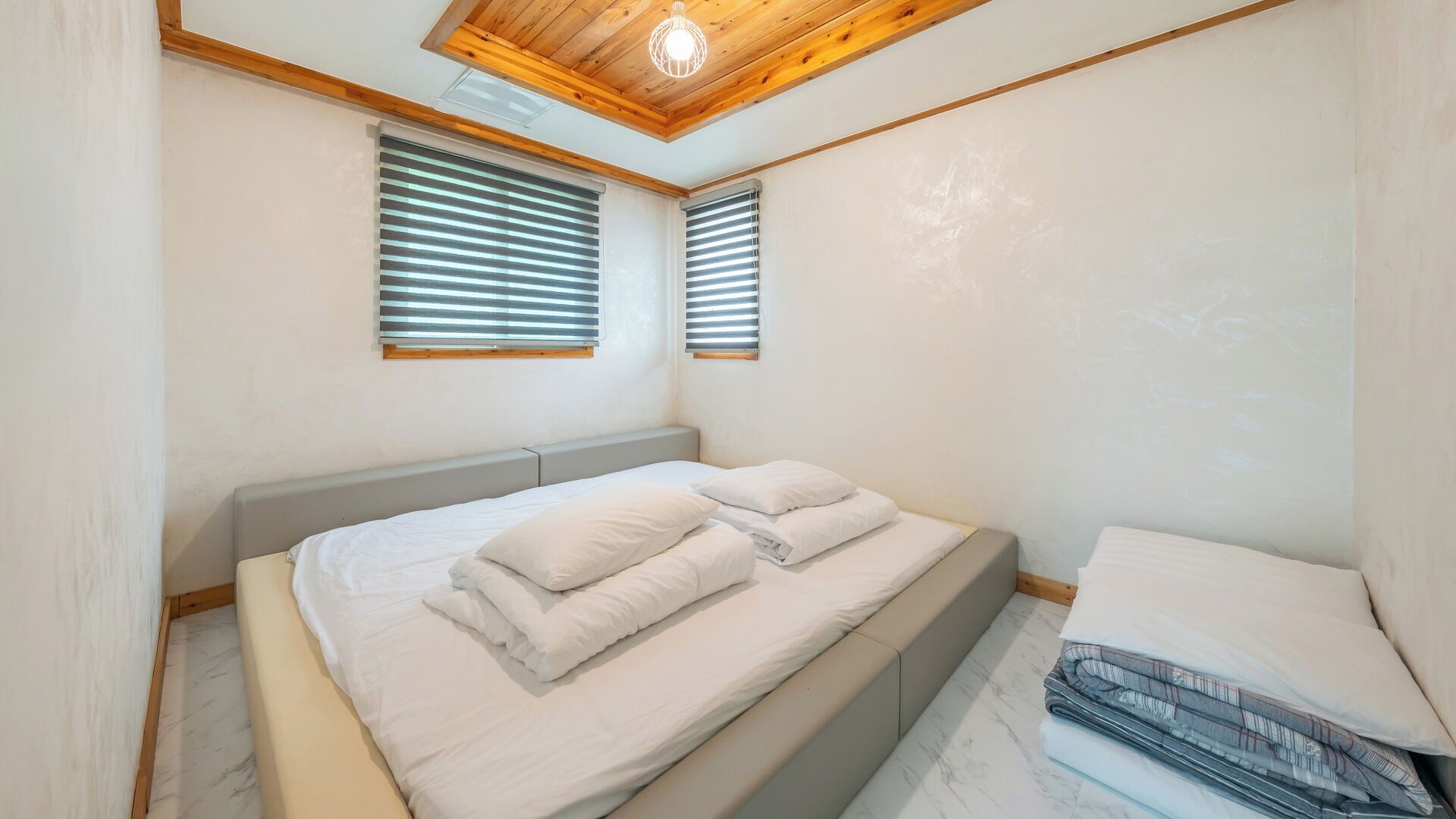A bedroom with a bed, two windows with blinds, a wooden ceiling, and a light fixture.