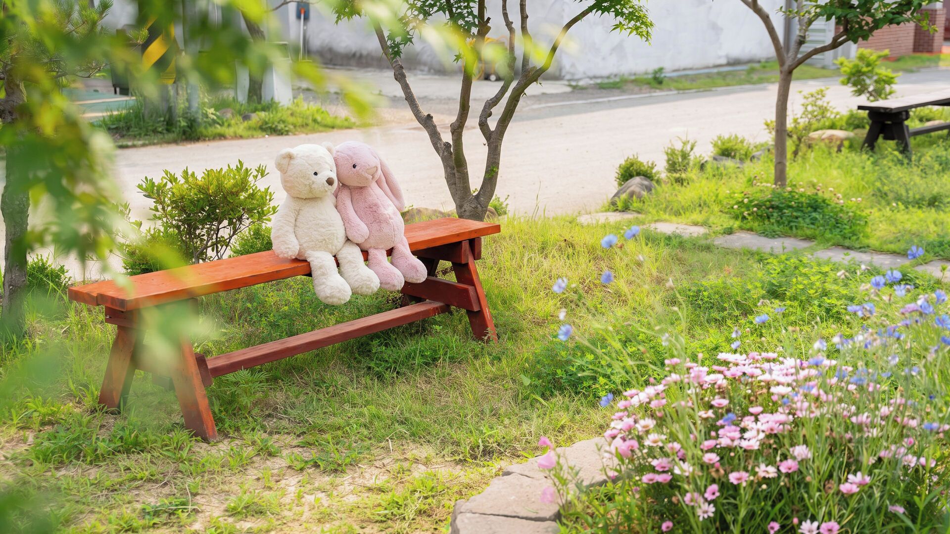 A wooden bench with two teddy bears, surrounded by grass and flowers.
