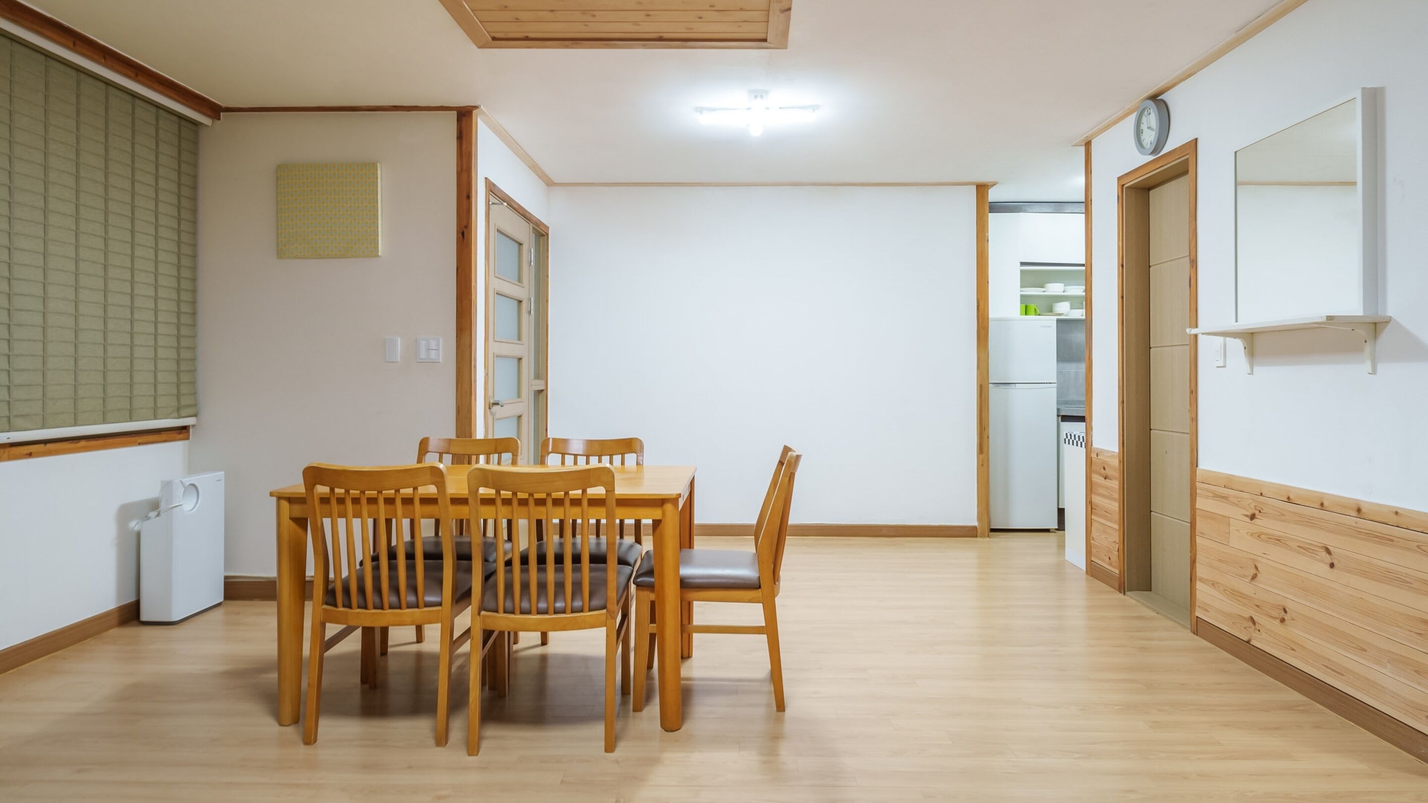 A spacious room with a wooden dining table and chairs, a small kitchen area, and a clock on the wall.
