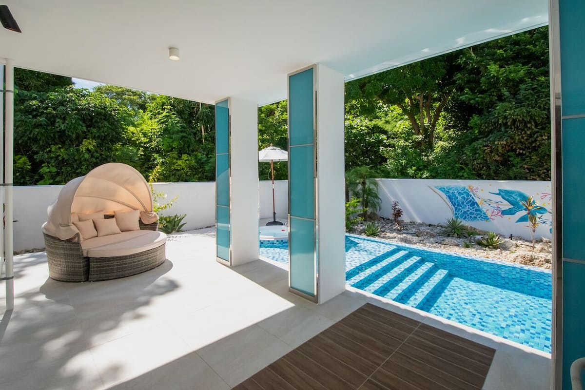 A modern outdoor patio with a pool, a cushioned seating area, and a view of a lush garden.