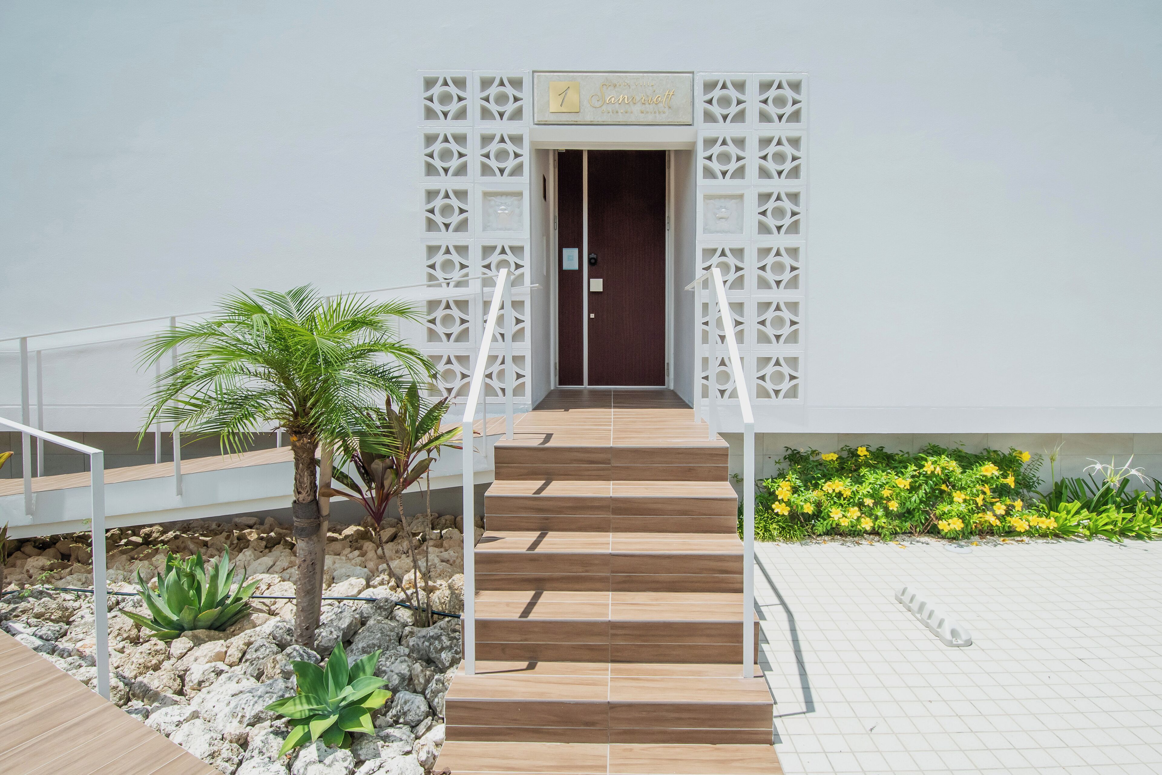 A modern building entrance with a wooden staircase leading to a dark door, surrounded by white lattice panels and a small garden with yellow flowers and a palm tree.