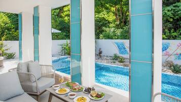 Un área de comedor junto a la piscina con una mesa dispuesta para dos, con vistas a una piscina y abundante follaje.