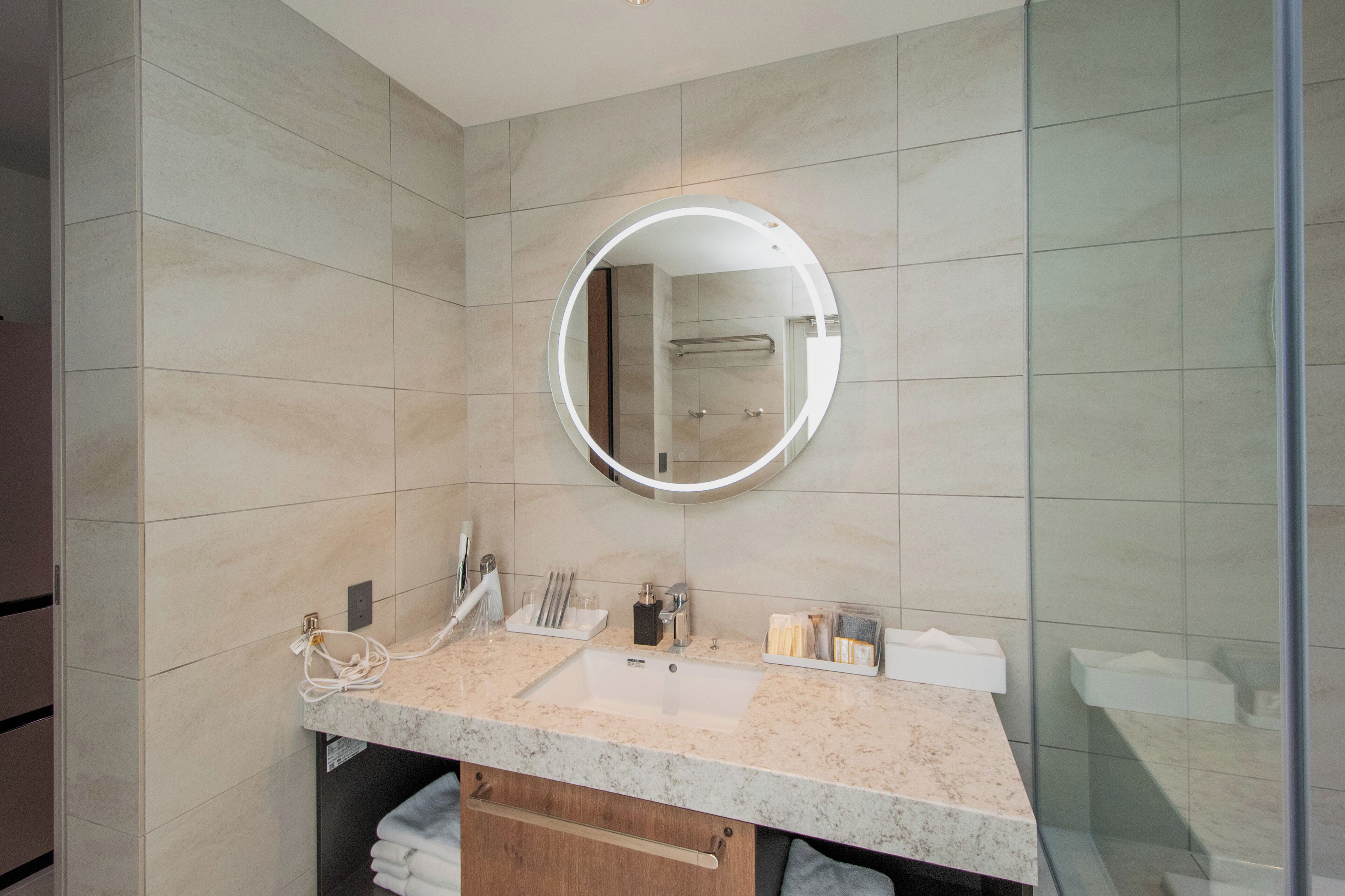 A modern bathroom with a marble countertop, a round mirror, and a glass shower enclosure.