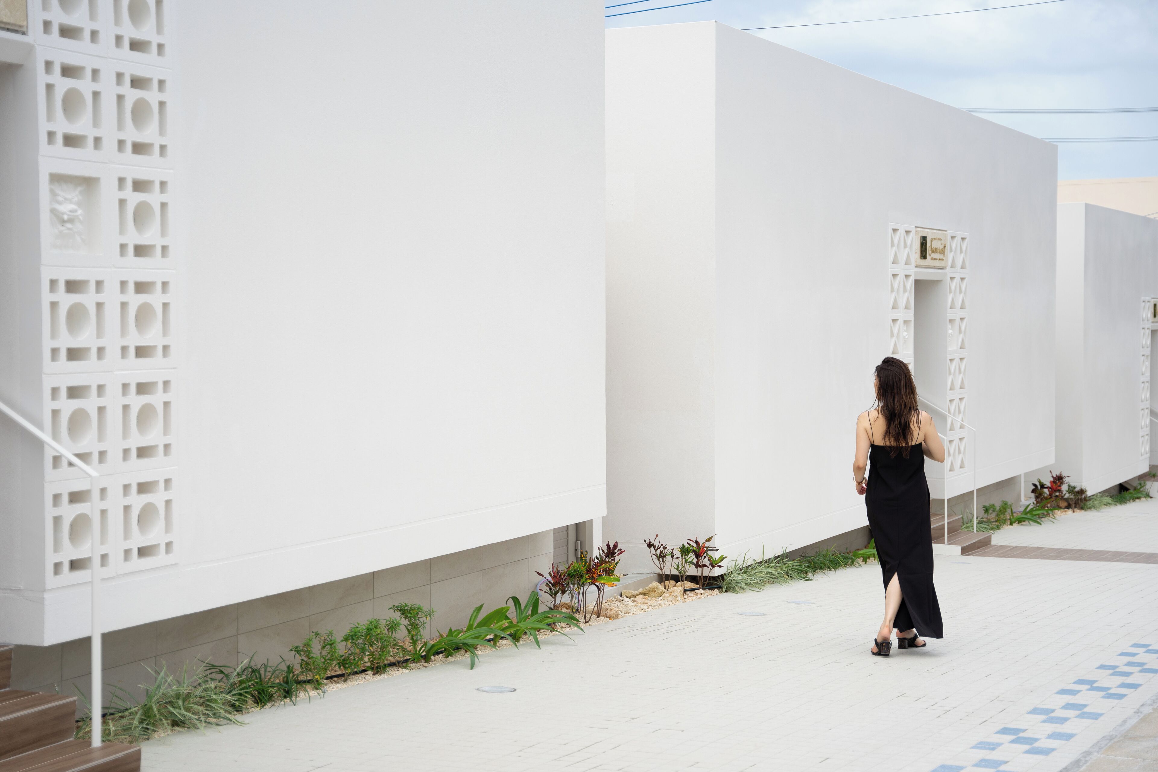 Une femme marche sur un chemin pavé de carreaux blancs formant un motif géométrique, à côté d’un bâtiment blanc dont les fenêtres sont décorées d’un motif en treillis.