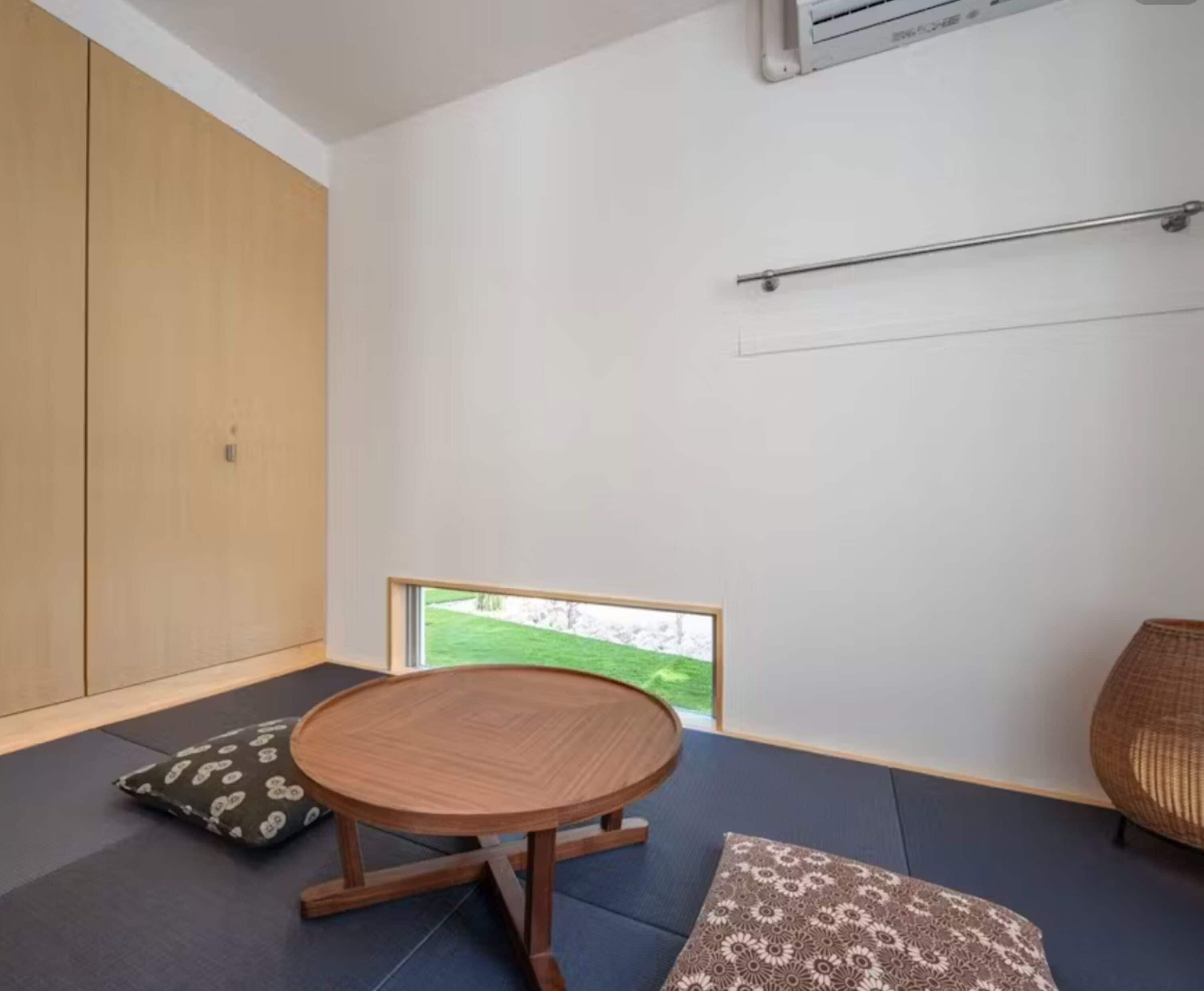 A room with a tatami mat floor, a round wooden table, a view of a green landscape, and a large wooden wardrobe.