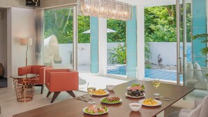 Un comedor moderno con una mesa de madera dispuesta para una comida, rodeado de asientos cómodos y ventanas amplias con vistas a una piscina y follaje.