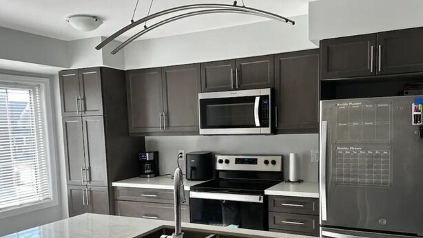 A modern kitchen with dark cabinets, stainless steel appliances, a built-in microwave, and a refrigerator with a weekly planner. - Cozy Whitby Retreat (Private 2 Bedroom Townhouse) (Whitby)