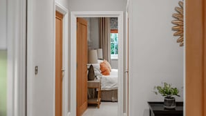 Ein Flur, der zu einem Schlafzimmer mit Bett, Nachttisch und einem mit Vorhängen versehenen Fenster führt.