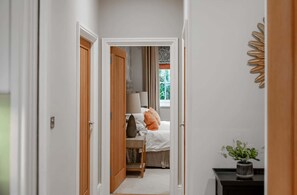 Un couloir mène à une chambre à coucher qui contient un lit, une table de chevet et une fenêtre avec des rideaux.