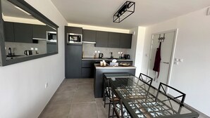 A modern kitchen with dark cabinets, a microwave, and a coffee machine. There is a glass dining table with chairs.