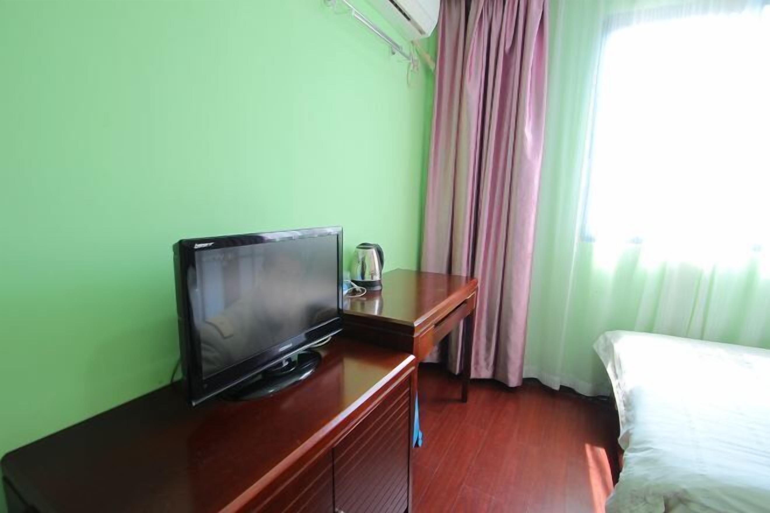 A hotel room with a bed, a TV on a wooden dresser, a window with curtains, and a wall-mounted air conditioner.