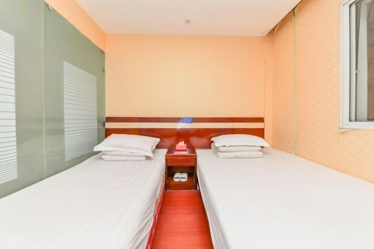 A hotel room with two single beds, a small table with a lamp, and a window.