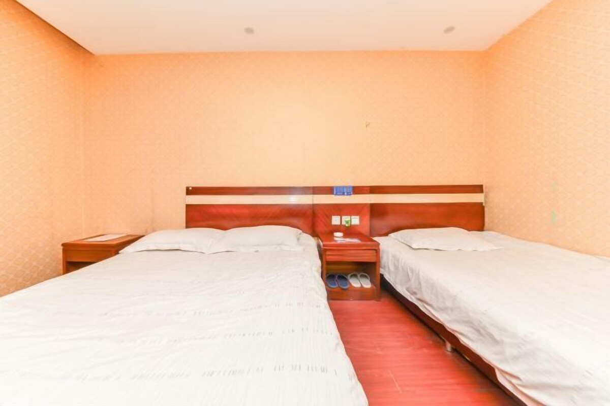 A hotel room with two single beds, a wooden headboard, a nightstand with a lamp, and a pair of slippers on the floor.