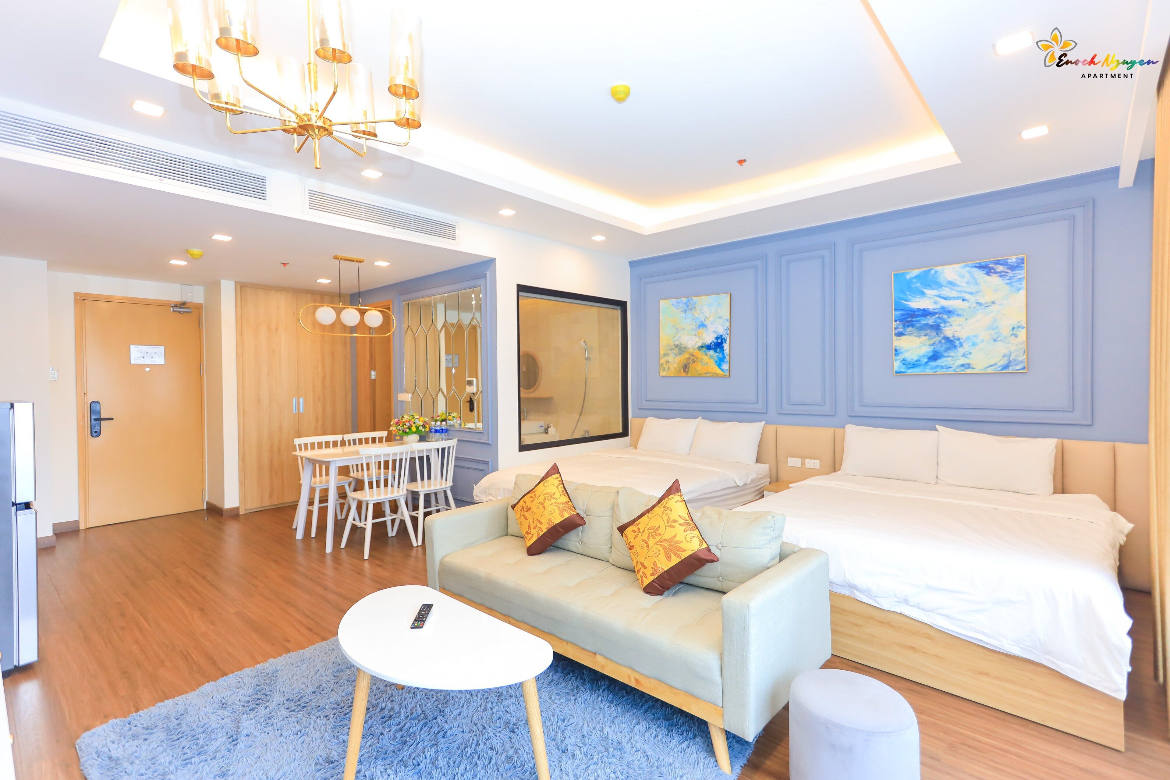 A modern hotel room with a large bed, a sofa, a dining table with chairs, and a chandelier.